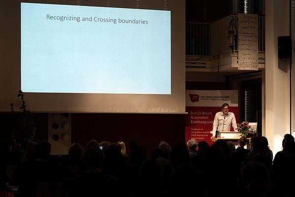 Laudator Stefan Köngeter am Redner_innenpult. Im Hintergrund eine Leinwand mit den Worten "Recognizing and Crossind Boundaries".
