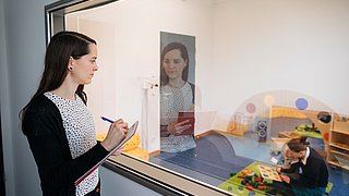 Eine Frau beobachtet durch eine verspiegelte Scheibe eine Mutter, die mit ihrer Tochter spielt.