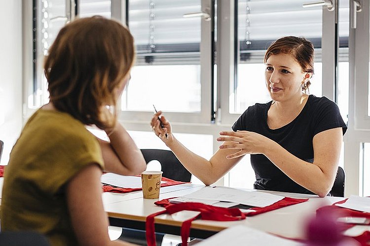Zwei Studentinnen sitzen sich an einem Tisch gegenüber. Eine Studentin erklärt der anderen etwas und gestikuliert mit ihren Armen.