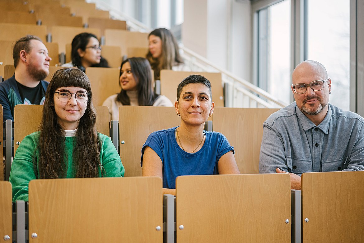 Studierende im Hörsaal. Drei schauen in die Kamera, die anderen unterhalten sich.