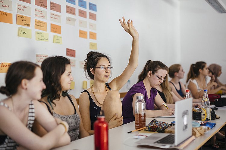 Studierende eines Seminars sitzen an einem langen Tisch nebeneinander. Eine Studentin meldet sich.