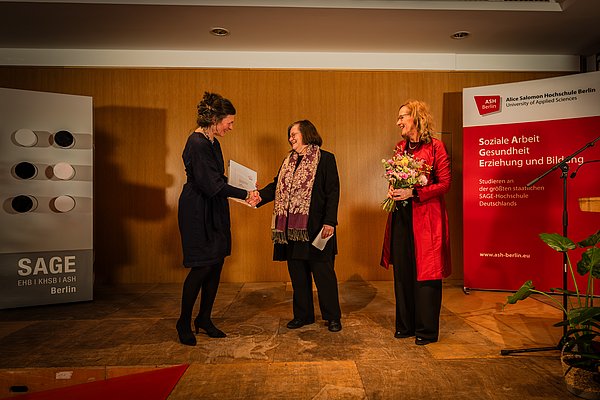 Bettina Völter und Anja Voss beglückwünschen Anna Csongor zum Alice Salomon Award und überreichen ihr eine Urkunde und einen Blumenstrauß.